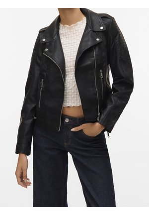 Vrouw draagt een zwarte leren bikerjack over een witte kanten crop top, gecombineerd met donkerblauwe spijkerbroek, hand in zak.