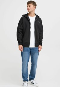 Zwarte zip-up hoodie met A.R.C. logo, gecombineerd met een wit T-shirt, lichtblauwe spijkerbroek en zwart-witte sneakers. Casual pasvorm en stijl.