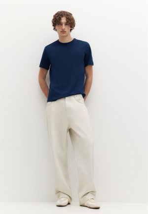 Jeune homme aux cheveux bouclés portant un t-shirt bleu marine, un pantalon blanc ample et des baskets blanches, debout contre un mur blanc avec les mains dans le dos.