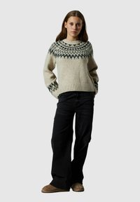 Jasno-beżowy sweter dzianinowy z okrągłym dekoltem oraz ciemnozielonym geometrycznym wzorem przy karczku. Połączony z czarnymi spodniami typu wide-leg i brązowymi kapciami.