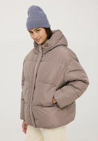 Giacca puffer marrone chiaro con cappuccio, chiusura con zip e due grandi tasche. Texture morbida con cuciture visibili e dettagli con cordino.