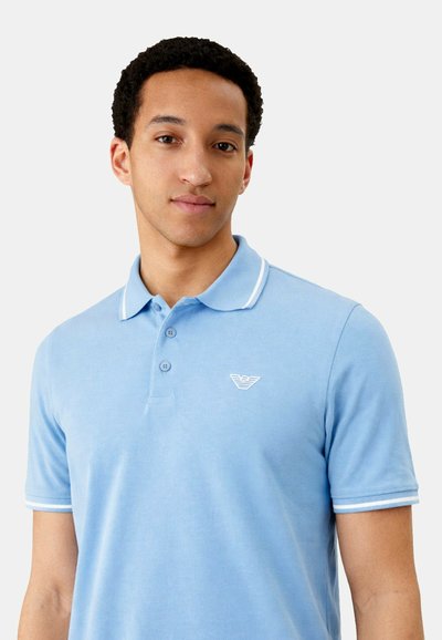 Jeune homme portant un polo bleu clair avec un col et des bords de manches blancs sur un fond uni.