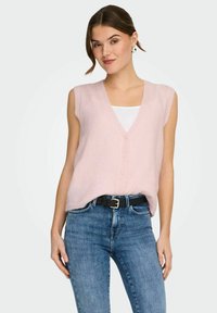 Jeune femme portant un gilet en tricot sans manches rose clair boutonné sur un haut blanc et un jean bleu avec une ceinture noire, debout devant un fond uni.