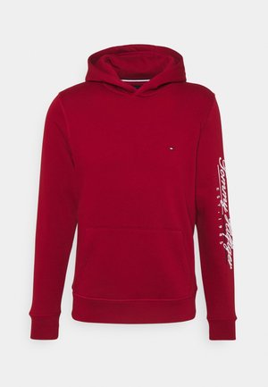 Czerwona bluza z kapturem z kieszenią z przodu, małe logo na piersi i biały napis „Tommy Hilfiger EST. 1985” na prawym rękawie.