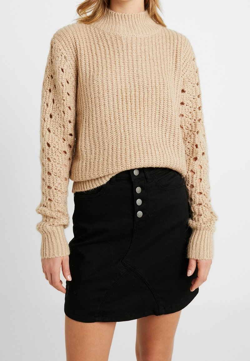 Femme portant un pull beige à gros mailles avec des manches texturées en ajouré et une mini-jupe noire taille haute à boutons sur le devant.