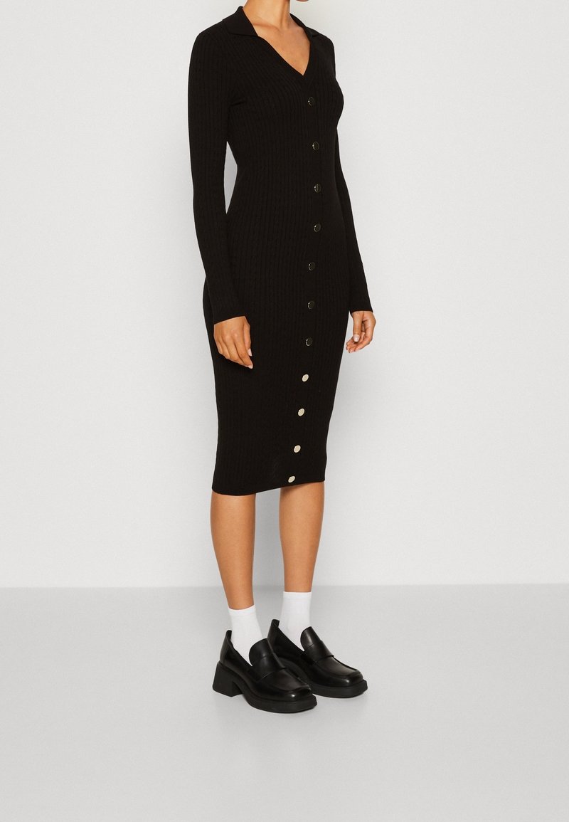 Femme portant une robe midi noire côtelée boutonnée avec manches longues, des mocassins noirs épais, et des chaussettes blanches aux chevilles sur un fond uni.