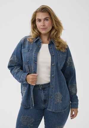 Blonde Frau trägt eine blaue Jeansjacke und Jeans mit Strass-Rosenmustern, posiert vor einem schlichten hellen Hintergrund.