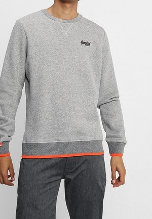 Mann trägt ein graues Rundhals-Sweatshirt mit dezenten Streifen, kleinem Brustlogo und orangefarbenen Akzenten an Bündchen und Saum, dazu eine dunkelgraue Hose.
