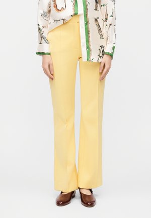 Osoba nosící žluté kalhoty s vysokým pasem, hnědé uzavřené boty a bílou košili se zeleným lemováním a zvířecími motivy.