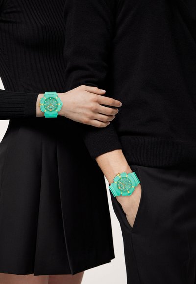 Due orologi da polso turchesi con dettagli dorati su abbigliamento nero. Un orologio ha un quadrante rotondo e un cinturino, caratterizzato da un design testurizzato.