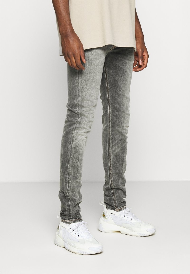 Pantalones vaqueros skinny grises descoloridos con una textura envejecida, combinados con zapatillas deportivas blancas gruesas. Los vaqueros cuentan con un sutil detalle de costura a lo largo de las costuras.