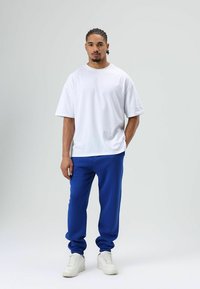 Witte oversized T-shirt gecombineerd met blauwe sweatpants en witte sneakers. Soepele stof met een ontspannen pasvorm, geribbelde manchetten en een rechte silhouet.