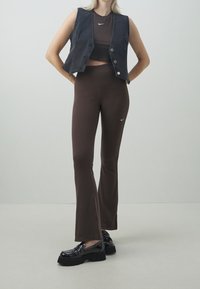 Brown flared pants and a dark denim vest, both featuring a small white Nike logo. The outfit is paired with black studded loafers.