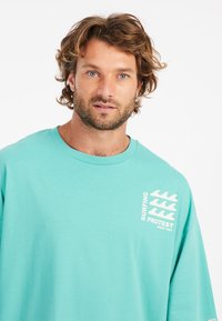 Tyrkysové oversized tričko s kulatým výstřihem a krátkými rukávy, s bílým textem a grafickými vlnami na přední straně.