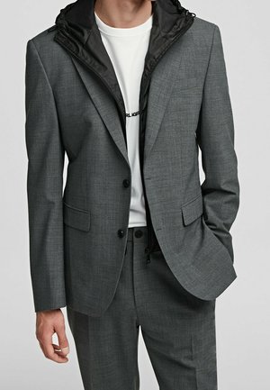 Mężczyzna ubrany w szary garnitur z czarną kurtką z kapturem pod spodem oraz białą koszulką z częściowo widocznym napisem.