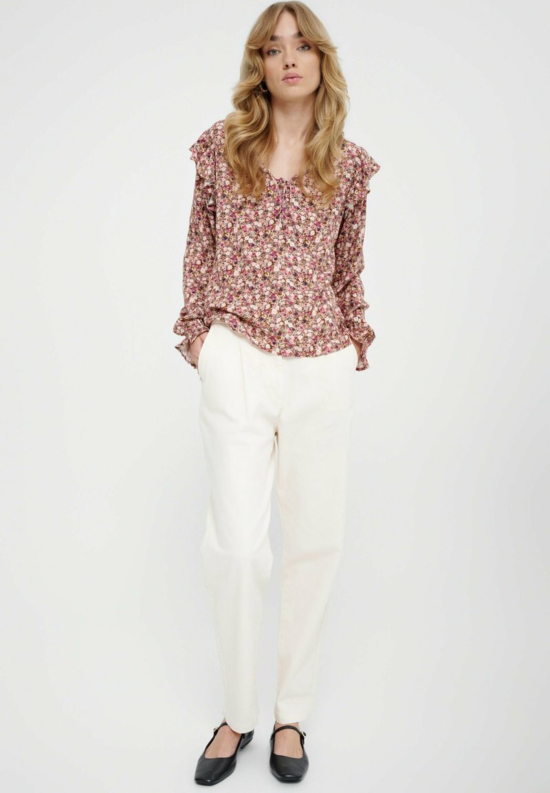 Kwiatowa bluzka z falbaniastymi rękawami, w odcieniach różu, bieli i zieleni; zestawiona z kremowymi spodniami w taperze i czarnymi balerinkami.