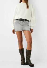 Mini jupe en denim gris associée à un gros pull blanc, ceinture noire à la taille et bottes brunes montantes avec des accents métalliques.