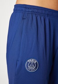 Pantalon de sport bleu en tissu lisse, équipé d'une taille élastique et d'un logo brodé rond du Paris Saint-Germain sur le côté.