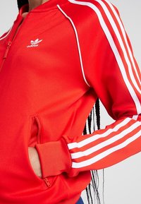 Červená bunda na zip s bílými akcentními proužky na rukávech, vyrobená z hladké látky, s bočními kapsami a malým logem na hrudi.