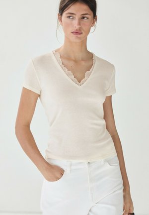 Femme portant un haut crème à manches courtes avec col en V et bordure en dentelle, ainsi qu'un pantalon blanc, debout avec une main dans la poche et regardant sur le côté.