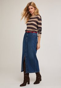 Gestreifter Strickpullover in Beige, Braun und Marineblau, kombiniert mit einem blauen Jeansrock mit Schlitz vorne, geschnallt mit einem roten Gürtel und braunen Stiefeln im Reptilmuster.