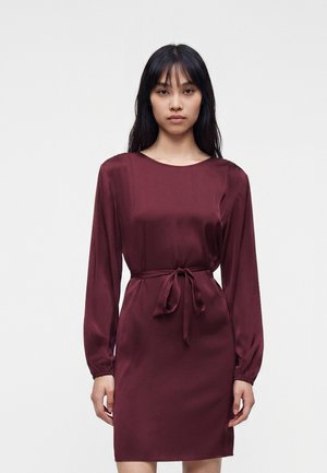 Donna con lunghi capelli neri che indossa un abito di raso bordeaux a maniche lunghe, lungo fino al ginocchio, con cintura annodata in vita, in piedi davanti a uno sfondo semplice.