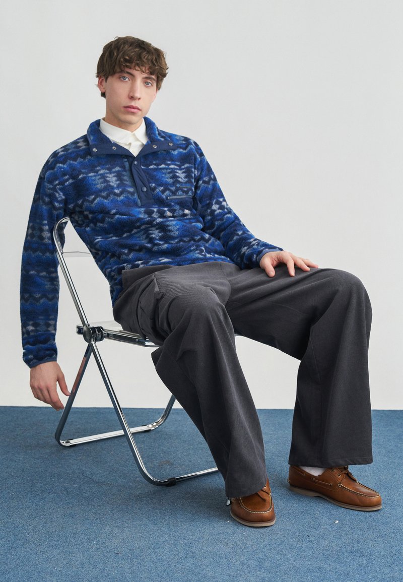 Νεαρός άνδρας καθισμένος σε πτυσσόμενη καρέκλα, φοράει μπλε φλις με σχέδια, λευκό πουκάμισο, γκρι παντελόνι με φαρδιές παντελόνες και καφέ δερμάτινα παπούτσια.