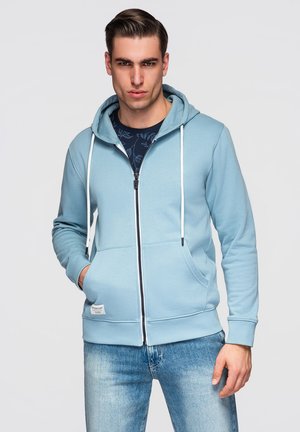 Jonge man met achterovergekamde haren, gekleed in een lichtblauwe hoodie met rits over een donkerblauw bloesje met bloemenprint en lichtblauwe spijkerbroek, handen in de zakken.