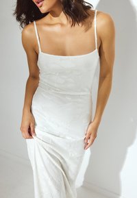 Robe blanche, ajustée, sans manches, avec un motif floral et un décolleté carré. Le tissu a un aspect doux et texturé.