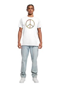 Hvid bomulds t-shirt med et stort, farverigt fredssymbol-tryk på forsiden. Modellen har lyseblå jeans og hvide sneakers på.
