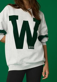 Jasnoszara bluza z dużym, ciemnozielonym logo "W", ściągaczami przy mankietach i talii. Etykieta z napisem "Wicked." Tło w jednolitym zielonym kolorze.