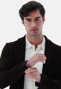 Blazer in corduroy marrone sopra una camicia bianca con righe verticali e colletto. Il blazer presenta bottoni sui polsini e una tasca.