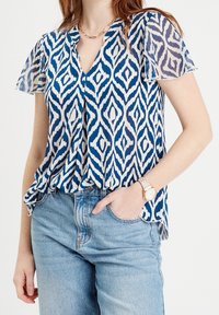 Blue and white patterned blouse with short sheer sleeves, V-neck design, and a loose fit. Paired with light blue denim jeans.