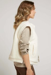 Gilet en polaire blanc sans manches, doté de poches latérales. Porté par-dessus un pull beige à manches longues. Modèle présenté de profil.