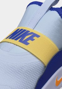 Λευκό και μπλε αθλητικό παπούτσι Nike με κίτρινο λουράκι στο επάνω μέρος, που φέρει μπλε γράμματα "NIKE" και μικρό πορτοκαλί σήμα Nike στο πέλμα.