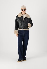 Veste cropped en cuir noir avec col et poignets en fausse fourrure beige, dotée de quincaillerie argentée et d'une fermeture éclair. Associée à un jean bleu foncé.