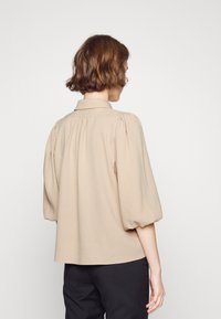 Beige blus med krage, puffade korta ärmar och smockdetalj på baksidan. Mjuk tyg och avslappnad passform. Bärs med svarta byxor.