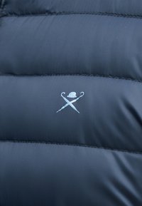 Σκούρο μπλε πουα ύφασμα με ένα μικρό φωτεινό μπλε λογότυπο που απεικονίζει ένα λευκό καπέλο μπούλερ πάνω από δύο σταυρωμένες ομπρέλες στο κέντρο της επιφάνειας.