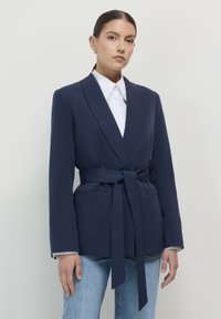 Donna che indossa un blazer blu navy con cintura sopra una camicia bianca con colletto e jeans blu chiaro, in piedi contro uno sfondo chiaro e uniforme.