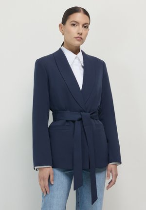 Frau, die ein marineblaues Blazer mit Gürtel über einem weißen Hemd und hellblauen Jeans trägt, vor einem schlichten, hellen Hintergrund steht.