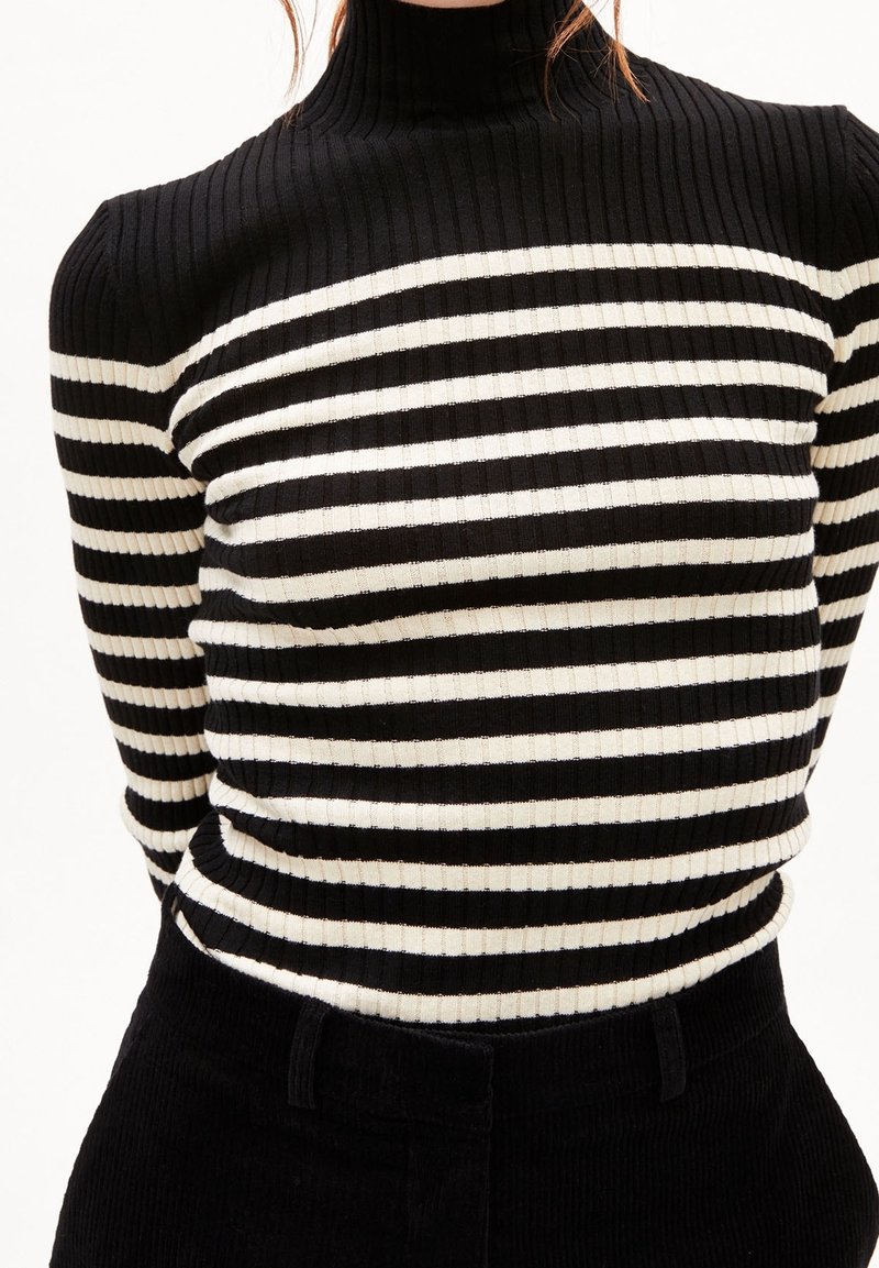 Personne portant un pull à col roulé côtelé rayé horizontal noir et blanc et un pantalon noir, coupé au niveau du cou et de la taille.