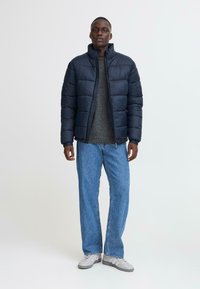Marineblå pufferjakke med høj krave, lynlåslukning og struktureret stof, parret med en grå sweater og løse blå jeans.