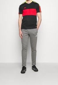 Zwarte sport T-shirt met een rode horizontale streep, gele accenten en grijze trainingsbroek met witte zijstrepen, gecombineerd met gestreepte zwarte schoenen.