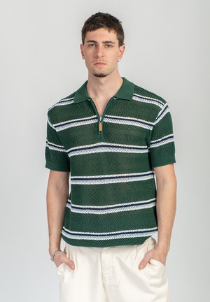 Homme portant un polo en maille vert à manches courtes avec des rayures blanches et marines, pantalon beige, debout devant un fond clair uni.