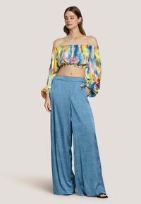 Top corto colorato fuori spalle con motivi vivaci; pantaloni larghi blu con una tessitura morbida. La modella posa su uno sfondo neutro.
