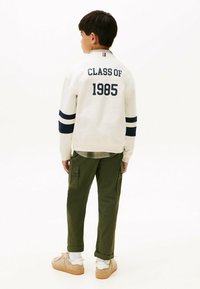 Witte pullover met "CLASS OF 1985" in marineblauw, gestreepte mouwen, gecombineerd met olijfgroene cargobroek en beige sneakers.