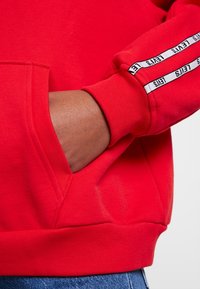 Red sweatshirt featuring a kangaroo pocket, ribbed cuffs, and a logo band along the sleeve. Material appears soft and textured.