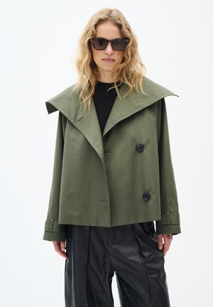 Veste cropped vert olive avec col large, double boutons et poignets en accent noir. Portée sur un haut noir et associée à un pantalon noir brillant.