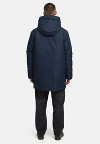 Jūras spēku garais mētelis ar kapuci, ar quilted tekstūru un taisnu griezumu. Apvienots ar tumšām biksēm un zābakiem, skatīts no aizmugures.