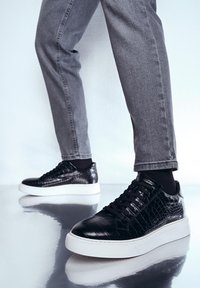 Sneaker in pelle nera con texture di coccodrillo, suola piatta bianca e lacci neri, abbinati a jeans skinny grigi.